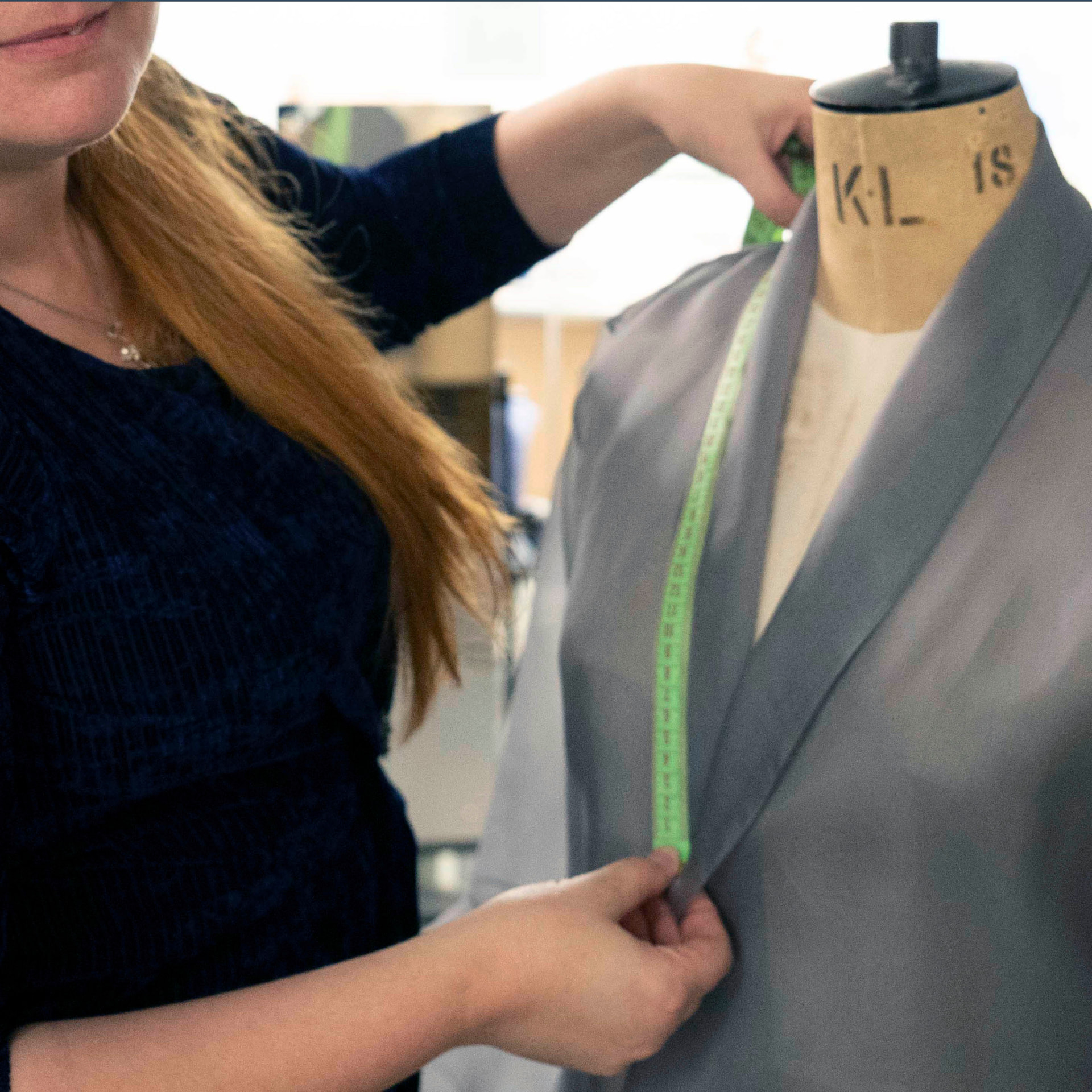 Seamstress measuring the neckline of a grey chiffon wrap garment on a dress form with a green tape measure at CMT Studio London