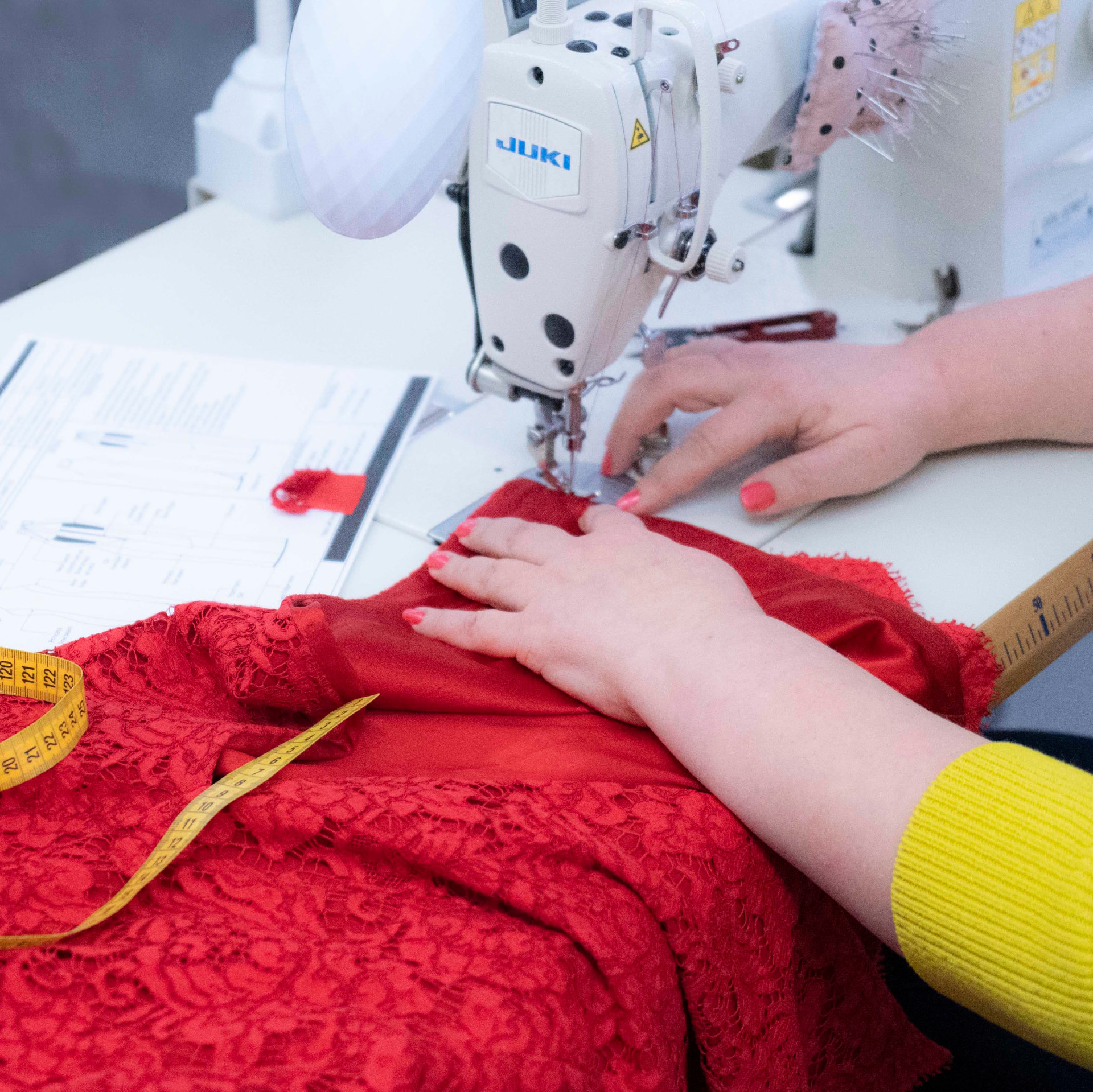 Machinist guiding red lace and satin fabric under a Juki sewing machine, with a tech pack and yellow tape measure beside it, at CMT Studio London