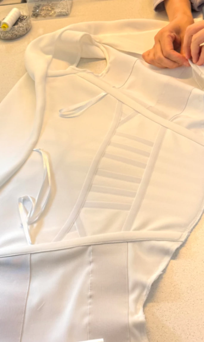 Hands hand-finishing a white bridal bodice with boning channels and ribbon ties on a worktable at CMT Studio London