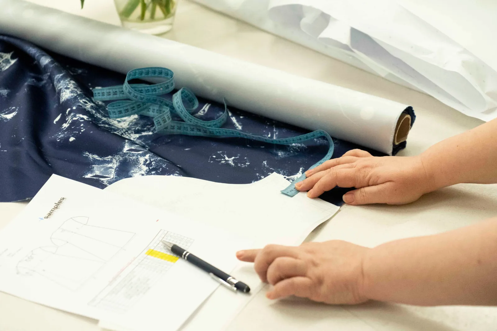 Hands smoothing a technical sketch beside navy printed fabric and a tape measure at CMT Studio London